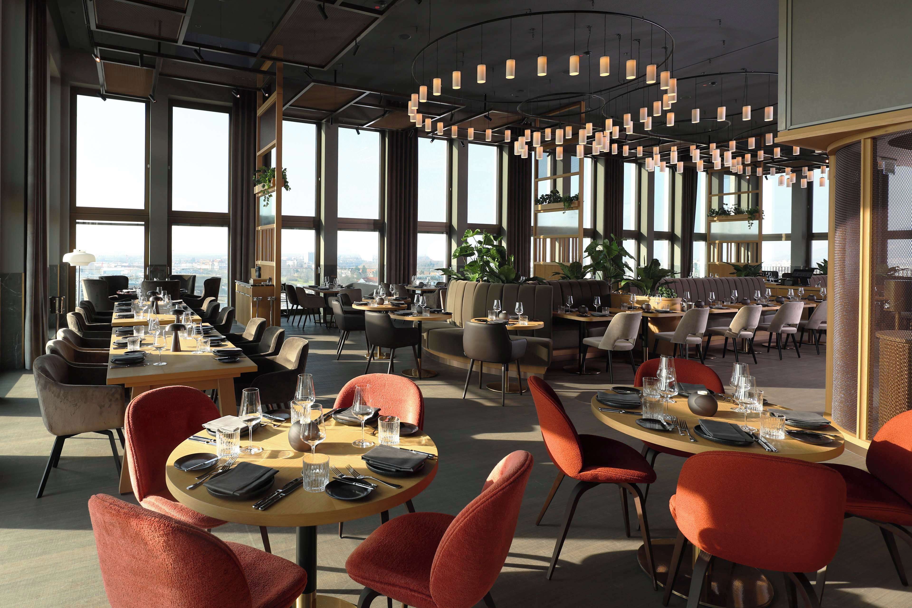 A modern dining area with multiple tables and chairs, large windows offering city views, and contemporary lighting fixtures.