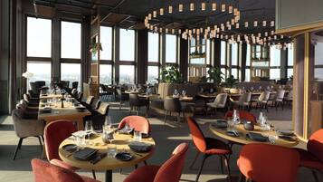 A modern dining area with multiple tables and chairs, large windows offering city views, and contemporary lighting fixtures.