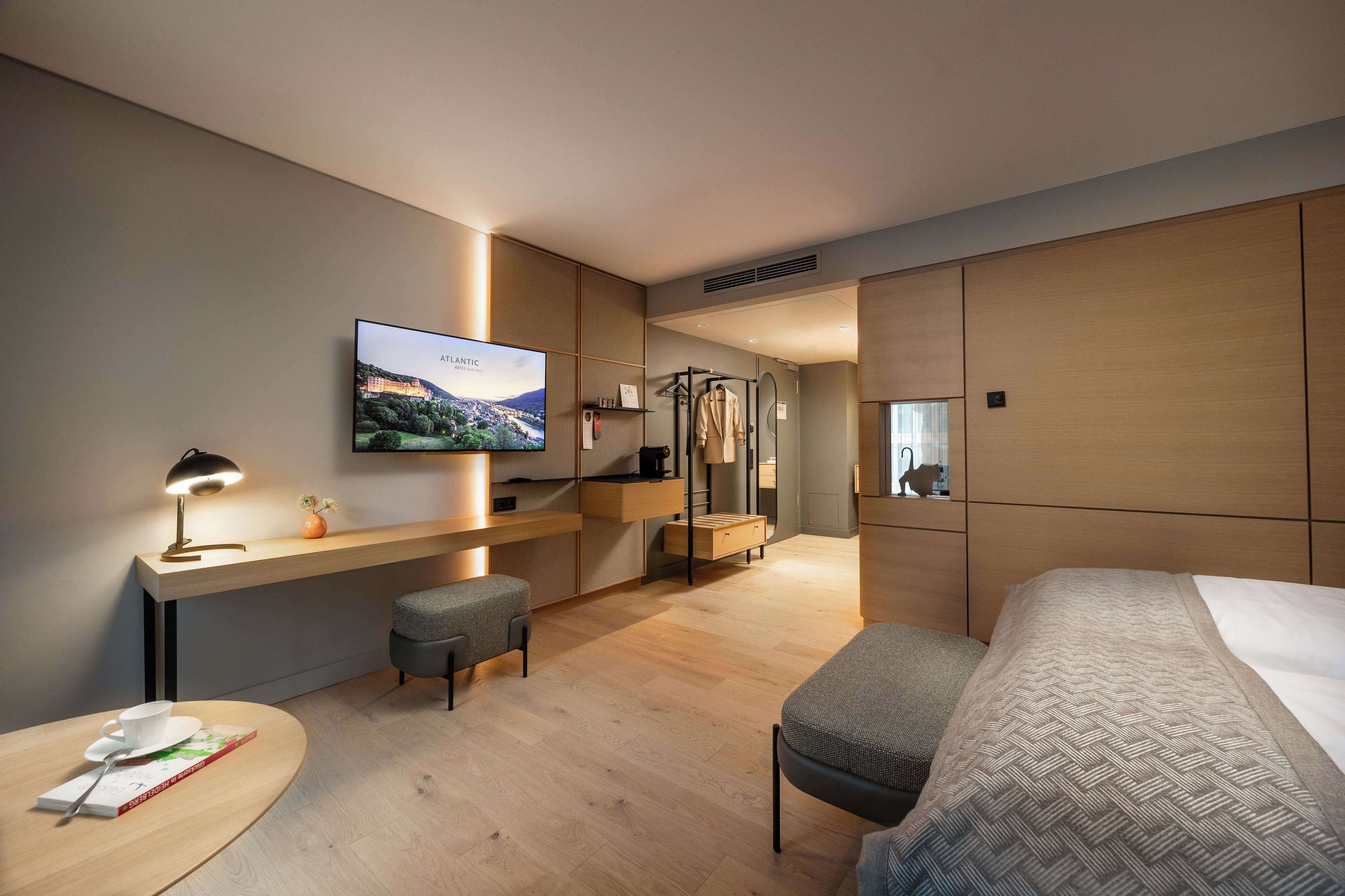 A modern hotel room with a large bed, a desk with a laptop, a TV mounted on the wall, and a small seating area.
