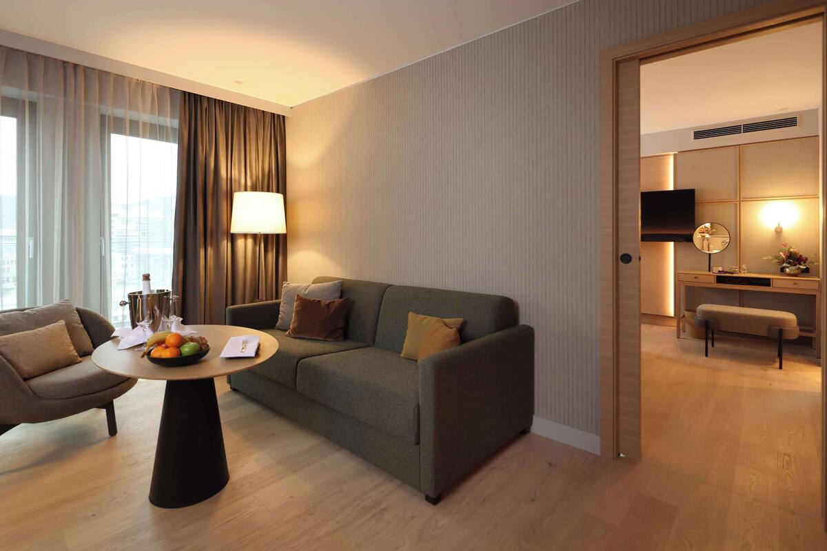 A modern hotel room with a sofa, armchair, and a round table with fruit.