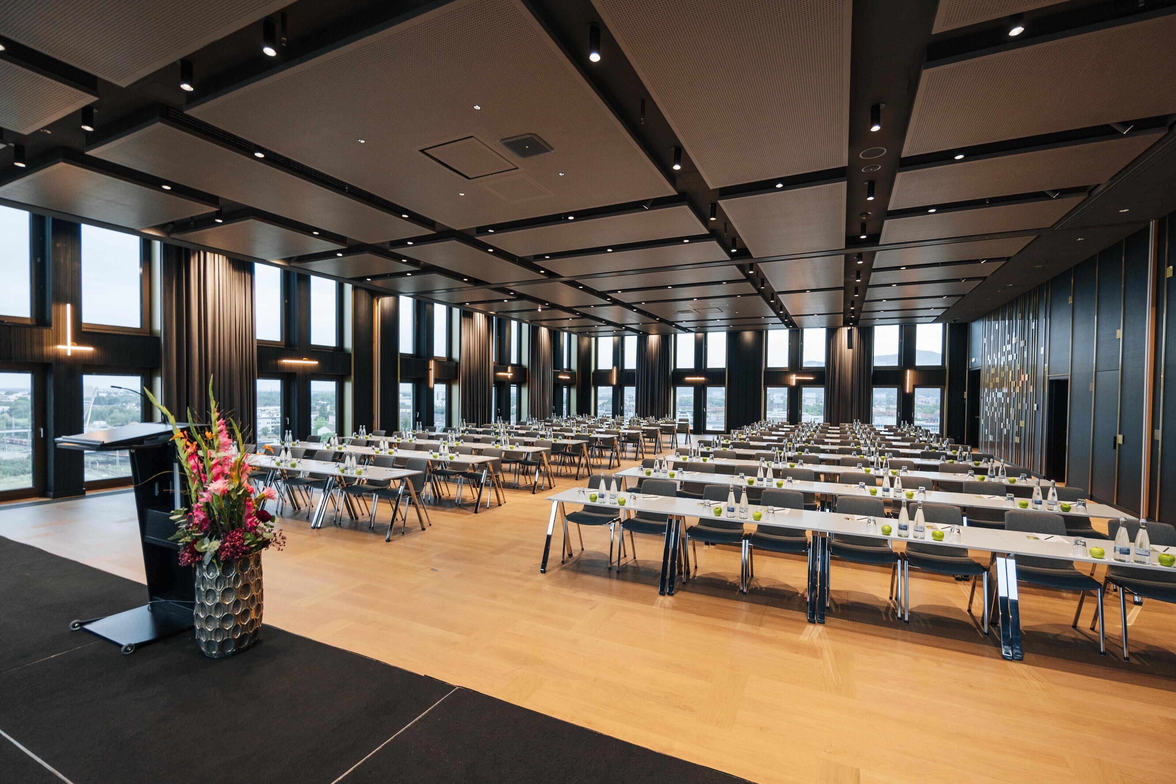Un salón de conferencias con varias mesas y sillas, ventanas amplias y un diseño moderno de techo.