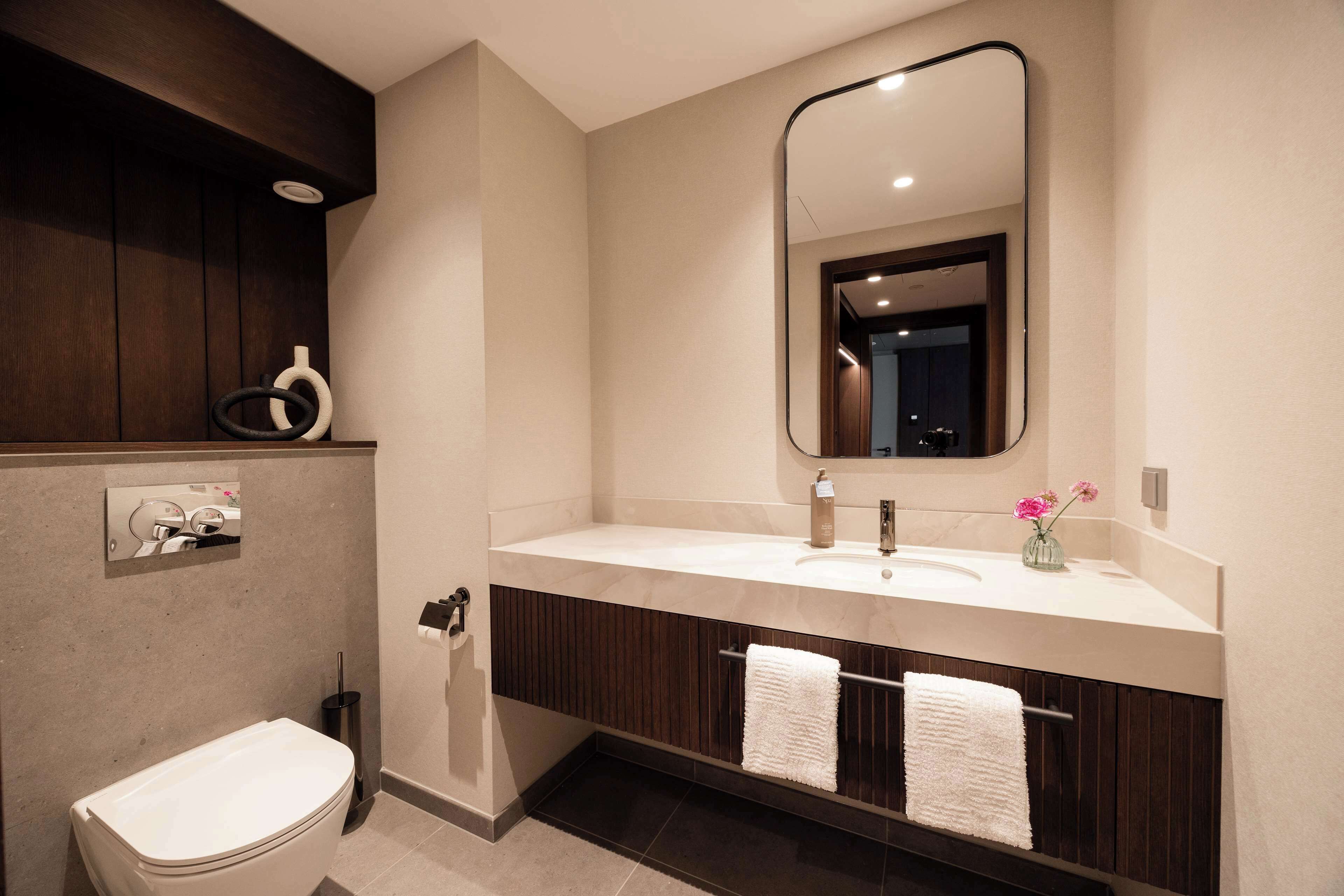 A modern bathroom with a white toilet, a large mirror, a vanity with a sink, and two towels hanging on a rack.
