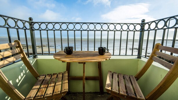 A balcony with a wooden table and two wooden chairs, overlooking a beach. - Home Away From Home (Newcastle)