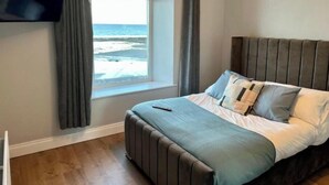 A bedroom with a bed, a TV, and a window with a view of the beach. - Home Away From Home (Newcastle)