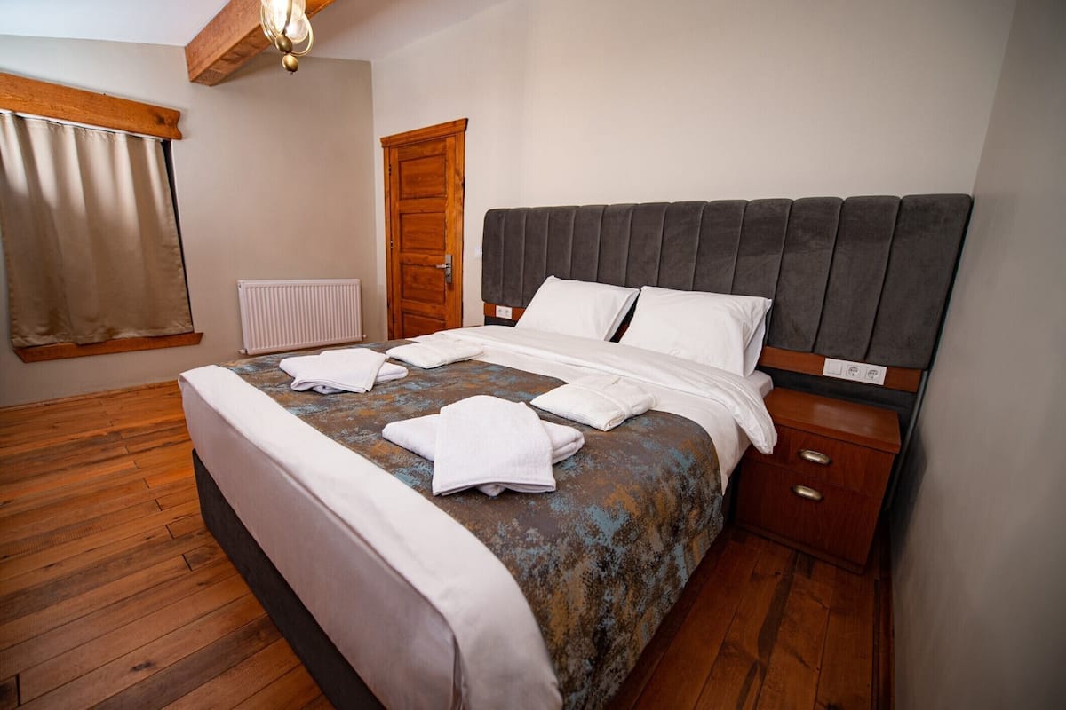 A neatly made bed with white linens and a grey headboard, a wooden nightstand with a drawer, a wooden floor, a window with curtains, and a radiator.