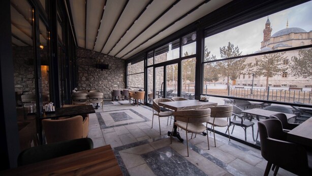 An interior with a stone wall, large windows, and a view of a mosque. - Ahıska Palas (Kars)