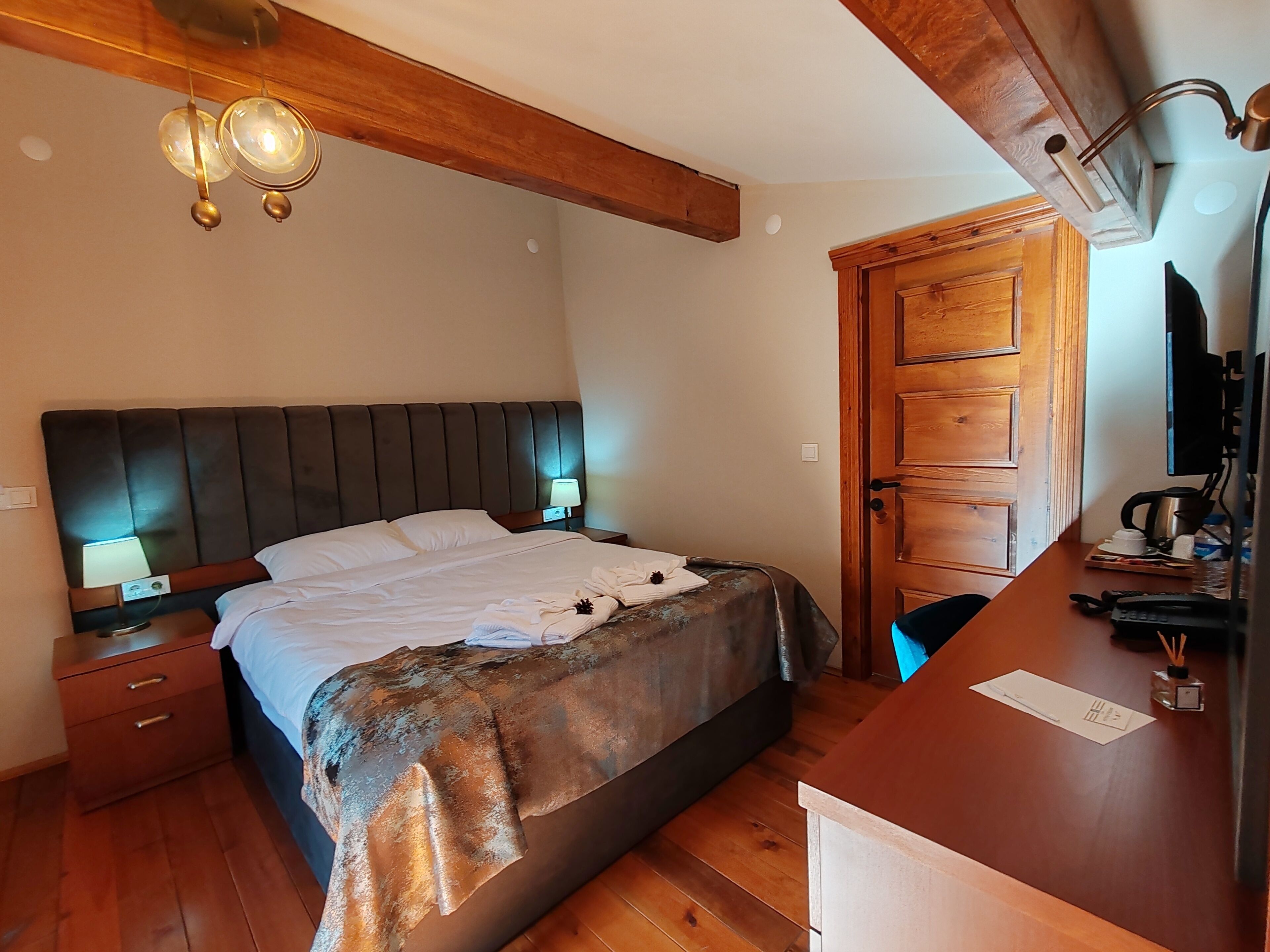 Un dormitorio con cabecera de madera, cama con sábanas blancas, dos lámparas de noche, un escritorio de madera con una computadora y una puerta de madera.