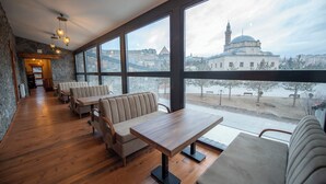 A restaurant with wooden tables and benches, large windows, and a view of a building with a dome and minaret. - Ahıska Palas (Kars)