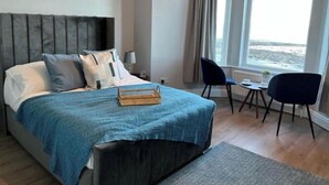 A bedroom with a bed, a bedside table, a chair, a small table, and a view of the sea. - Apartment - Ground Floor (Newcastle)