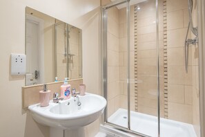Un baño con lavamanos blanco, espejo, cabina de ducha de vidrio y azulejos color beige.