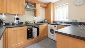 A modern kitchen with wooden cabinets, a stainless steel range hood, a built-in oven, a washing machine, and a window with blinds. - Apartment 8 (Worksop)