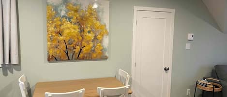 A dining area with a wooden table and white chairs, a white door, and a painting of yellow trees on the wall.