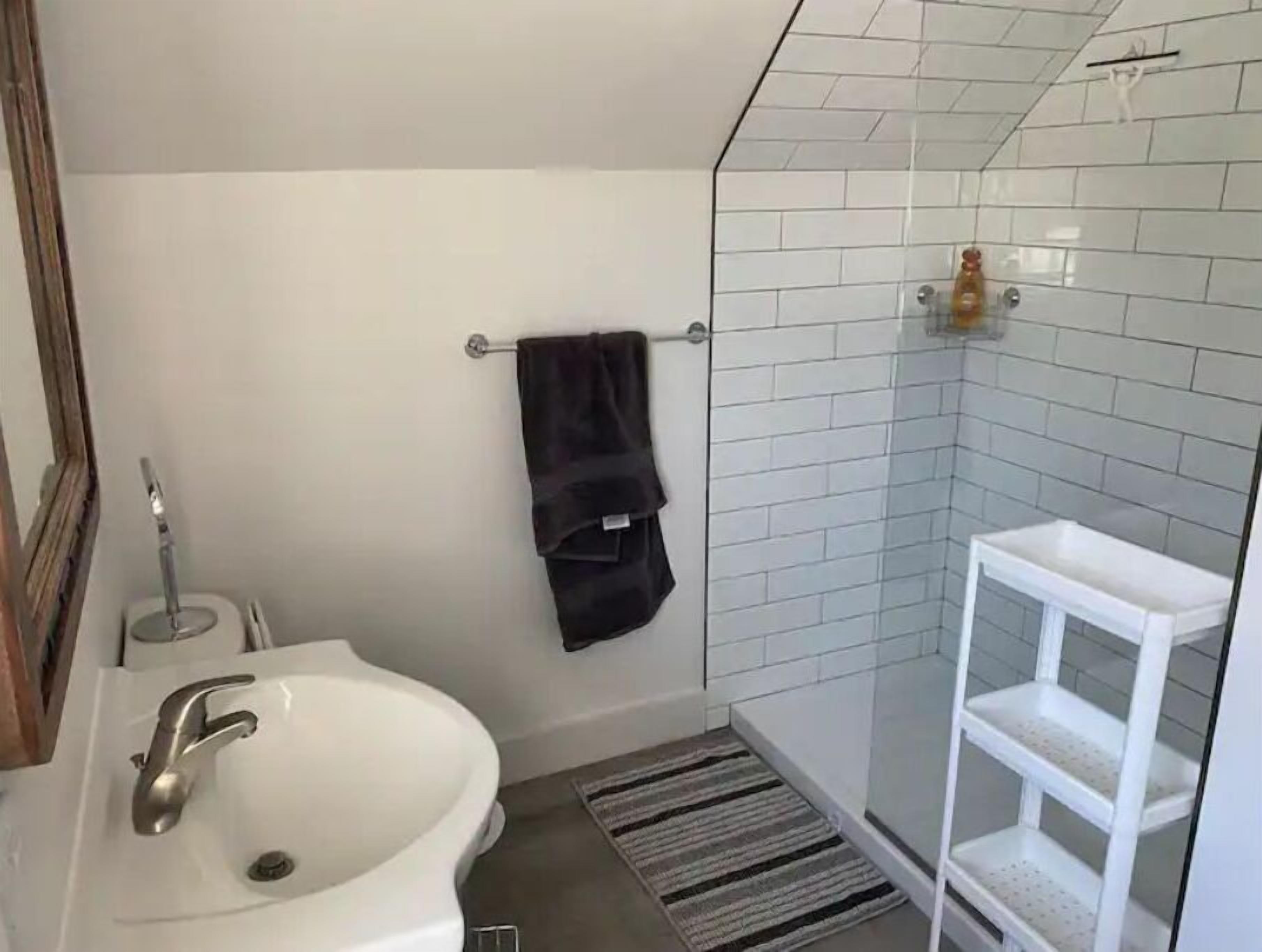 Un baño con un lavamanos blanco, un perchero con una toalla negra, un box de ducha de vidrio y un estante blanco.