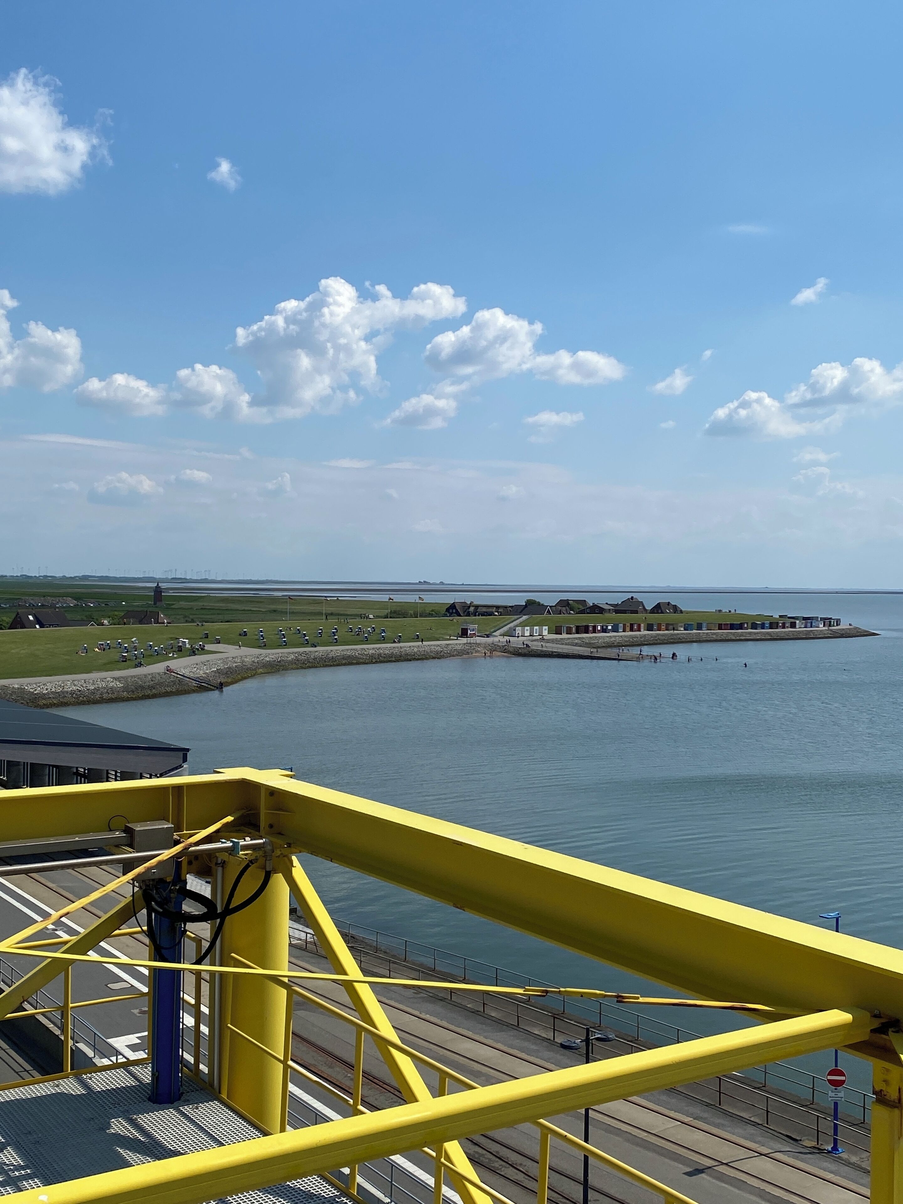 Blick auf die Küste von einer erhöhten Position, mit einer gelben Metallkonstruktion, ruhiger See und einer fernen Küstenlinie mit Gebäuden und Grünflächen.
