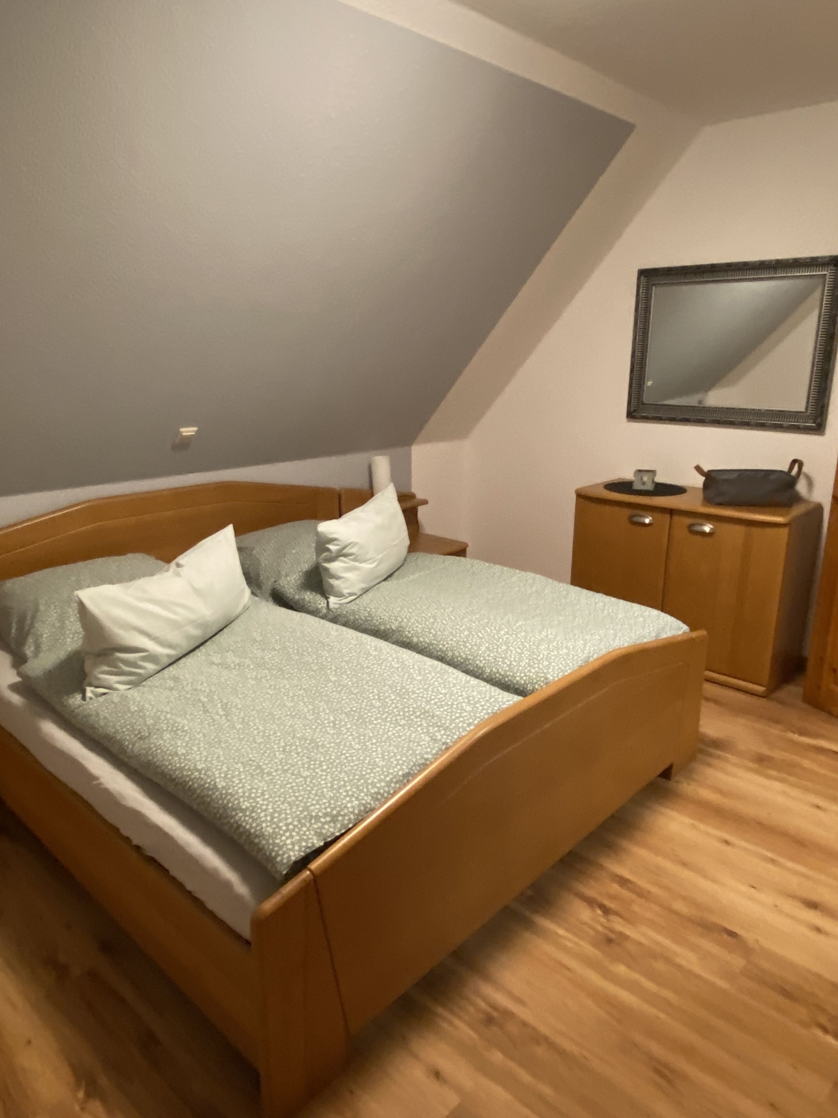Een slaapkamer met een houten bed, twee kussens, een nachtkastje, een dressoir en een spiegel aan de muur.