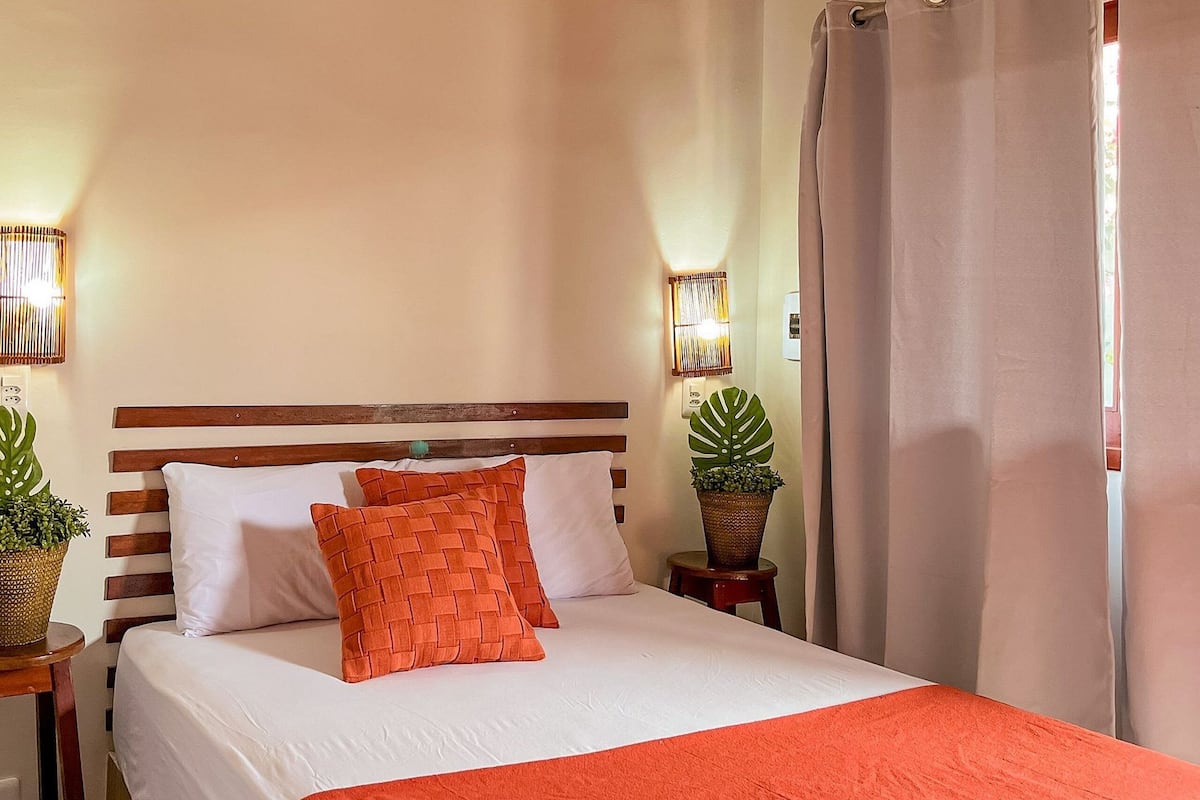 A neatly made bed with white and orange bedding, two bedside tables with lamps, and a potted plant on one table.