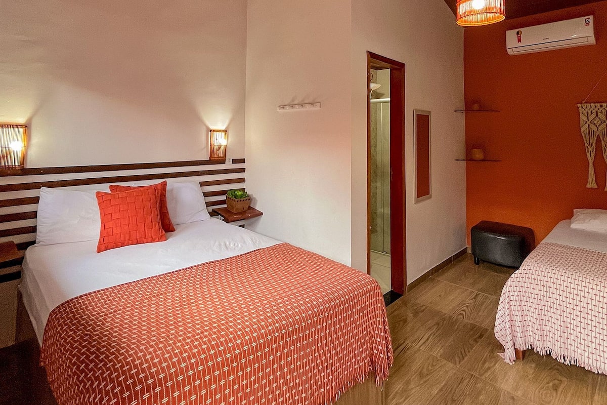A hotel room with a large bed, a smaller bed, a nightstand with a lamp, a mirror, and an air conditioning unit on the wall.