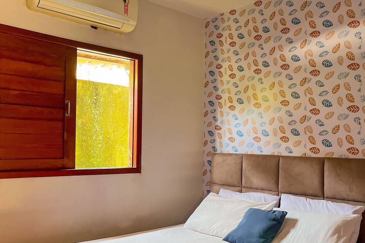 A bedroom with a bed, a window, an air conditioner, and patterned wallpaper.
