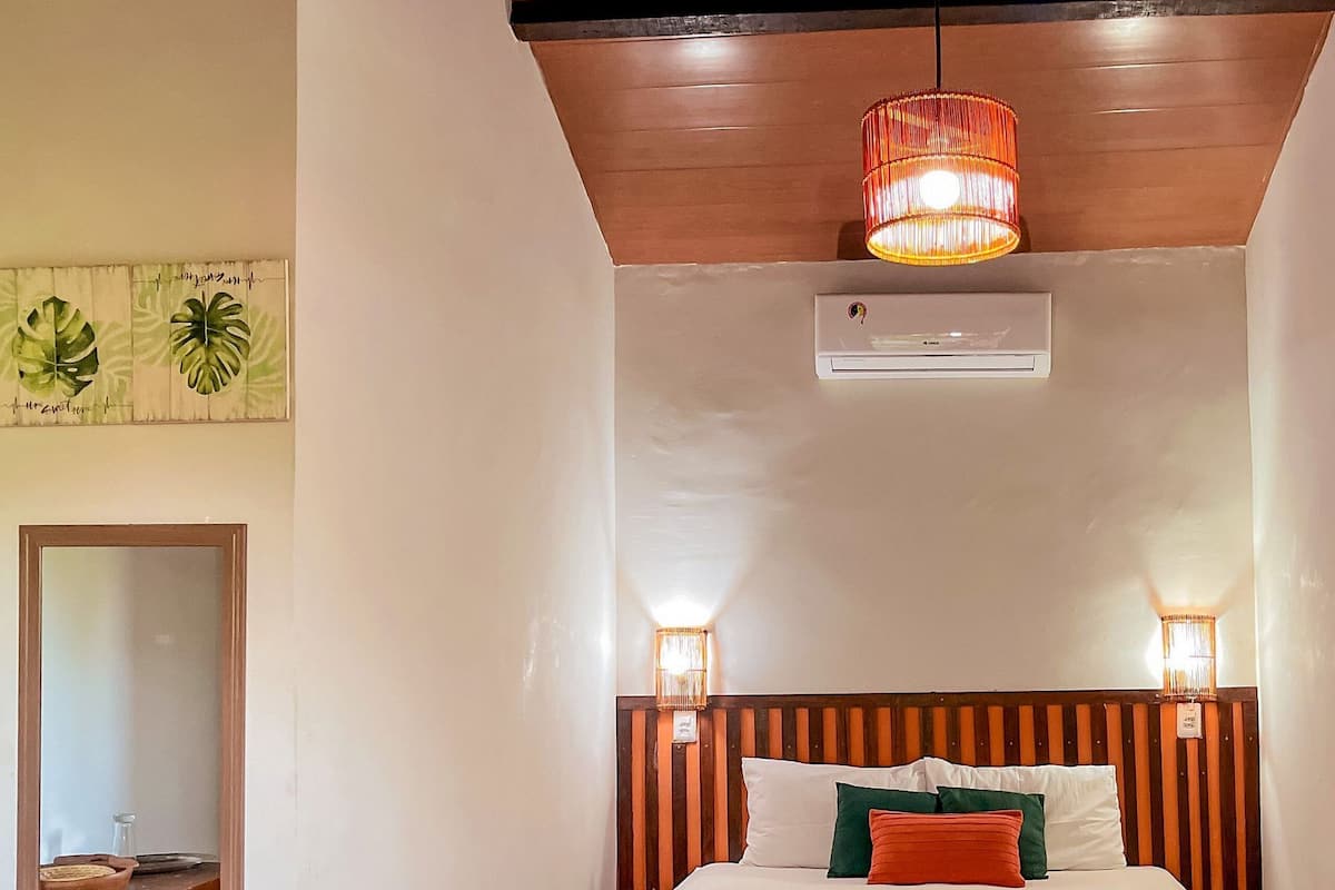 A bedroom with a wooden headboard, a bed with a pink bedspread, two bedside lamps, a mirror, and a small table with a bowl and a bottle.