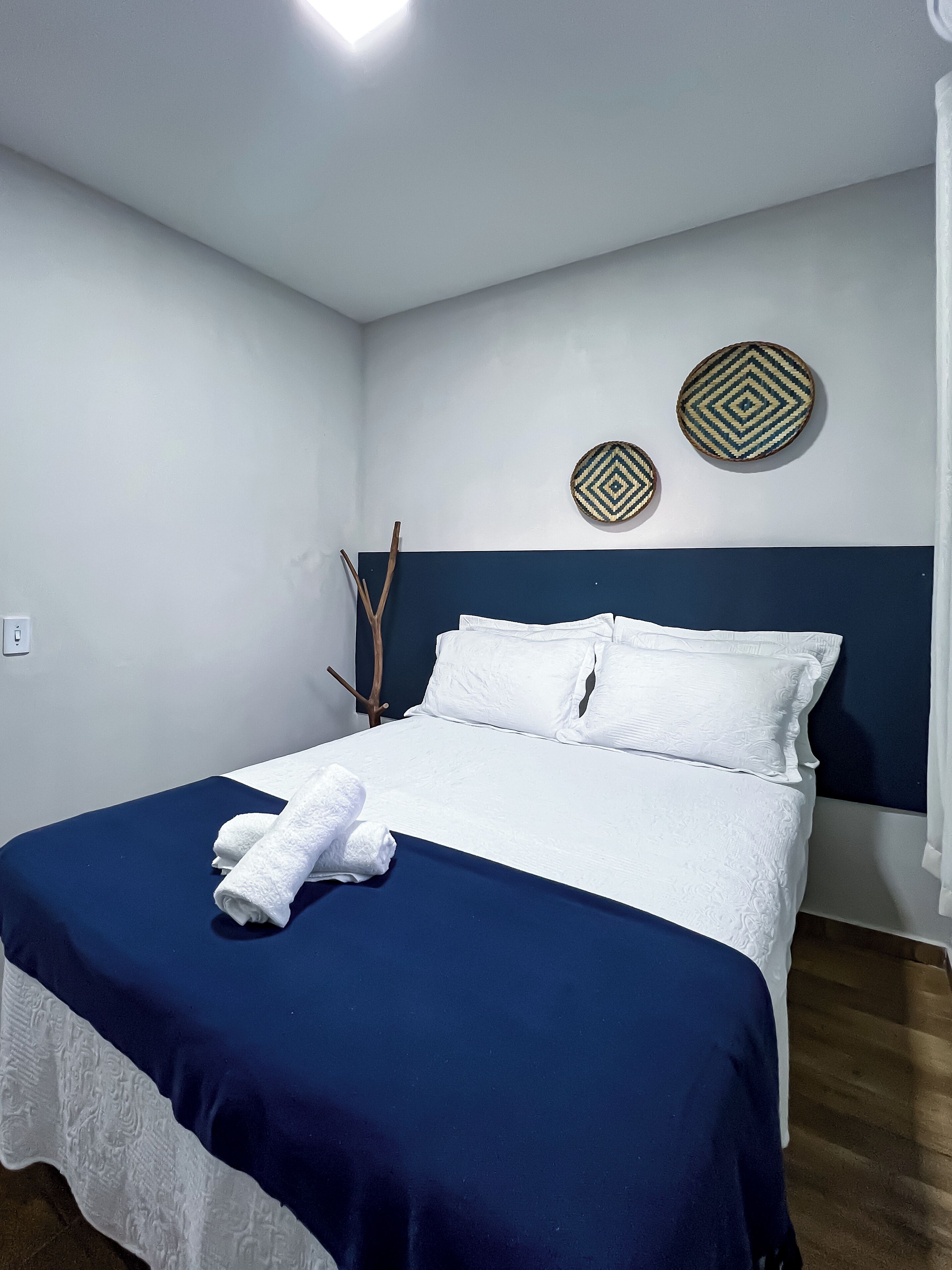 Un lit bien fait, avec des draps blancs et une couvre-lit bleue, deux décorations murales tissées et une branche en bois accrochée au mur.