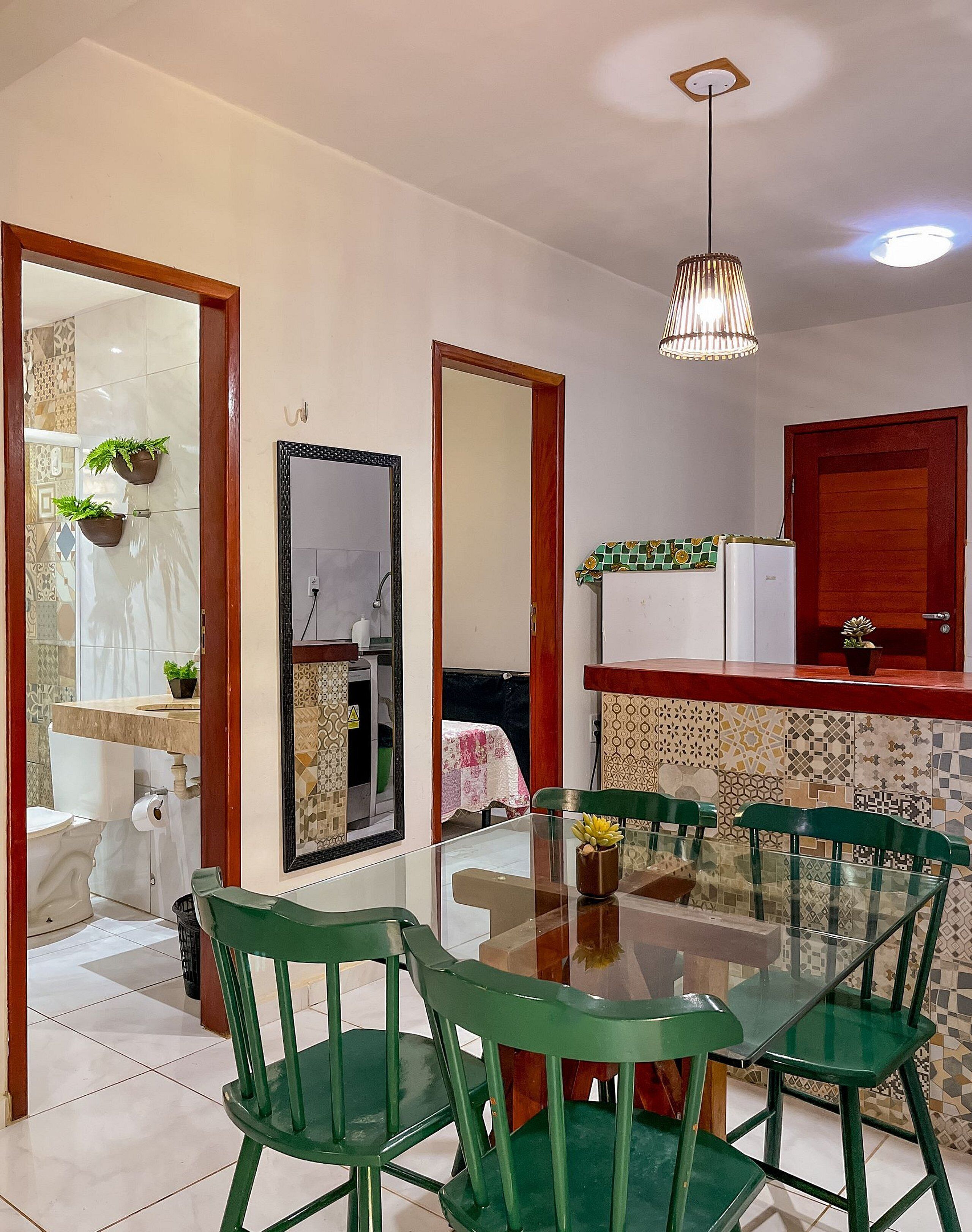 A dining area with a glass table and green chairs, a kitchen with a refrigerator and tiled backsplash, and a bathroom with a toilet and mirror.