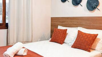 A neatly made bed with a red blanket and two pillows, a wall-mounted air conditioner, and three decorative hats on the wall.