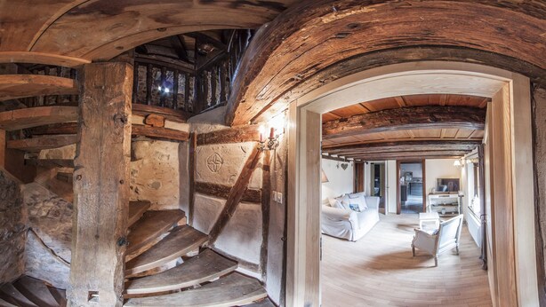 A spiral staircase in a rustic stone building with a wooden ceiling and a view into a modern living area with a sofa and a TV. - Cottage 'BRAND (Turckheim)