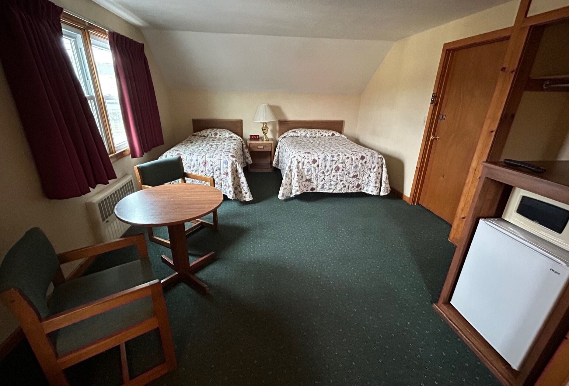 A small, cozy room with two single beds, a wooden table, a chair, and a small white refrigerator.
