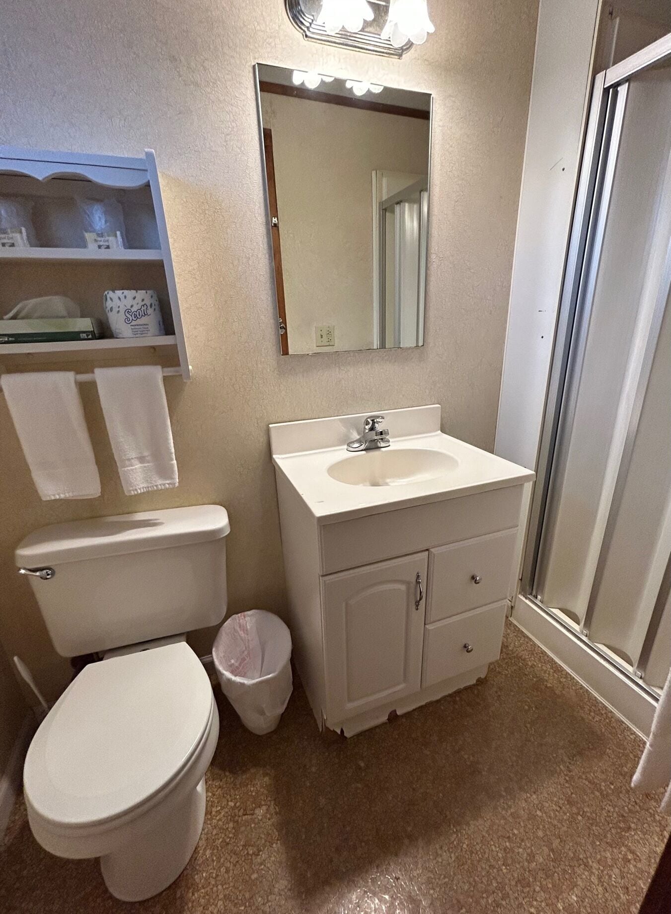 A bathroom with a toilet, sink, and mirror. There is a shelf with towels and a trash can.