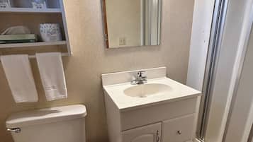 A bathroom with a toilet, sink, and mirror. There is a shelf with towels and a trash can.