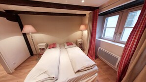 A bedroom with a large bed, two bedside tables, two lamps, a window with curtains, and a radiator. - Gîte 'WINSTUB (Turckheim)
