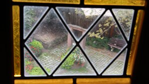 A window with a geometric pattern, through which a garden with a wooden structure and potted plants is visible. - Gîte 'WINSTUB (Turckheim)
