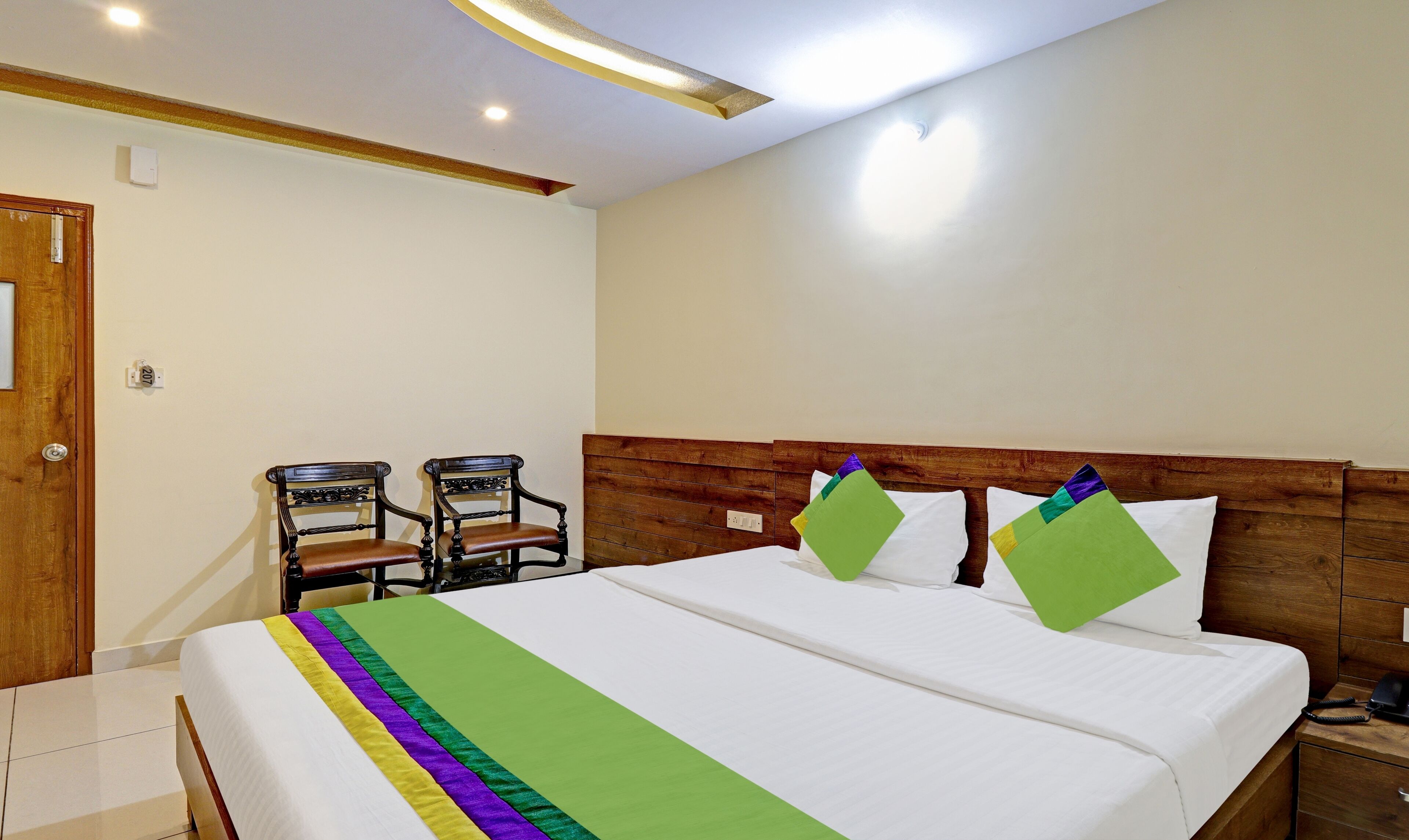 A hotel room with a large bed, two chairs, a wooden headboard, and a bedside telephone.