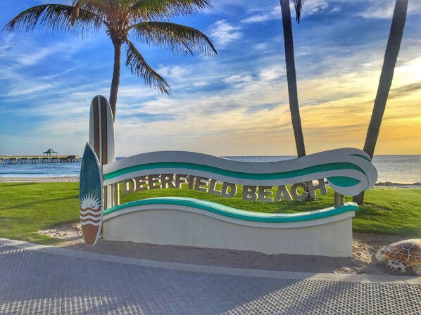 Un panneau de signalisation sur la plage avec l’inscription « Deerfield Beach » et une planche de surf.