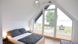 A bedroom with a large window, a bed, a bedside table with a lamp, and a hanging light fixture.