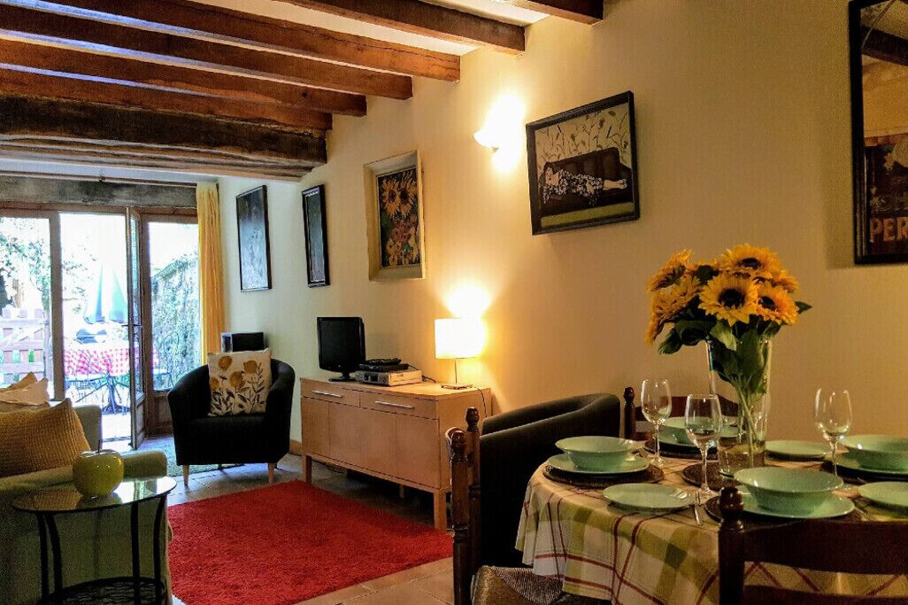 Un salon chaleureux avec un coin repas, des poutres en bois, une télévision et une table dressée pour deux personnes, agrémentée d’un vase de tournesols.