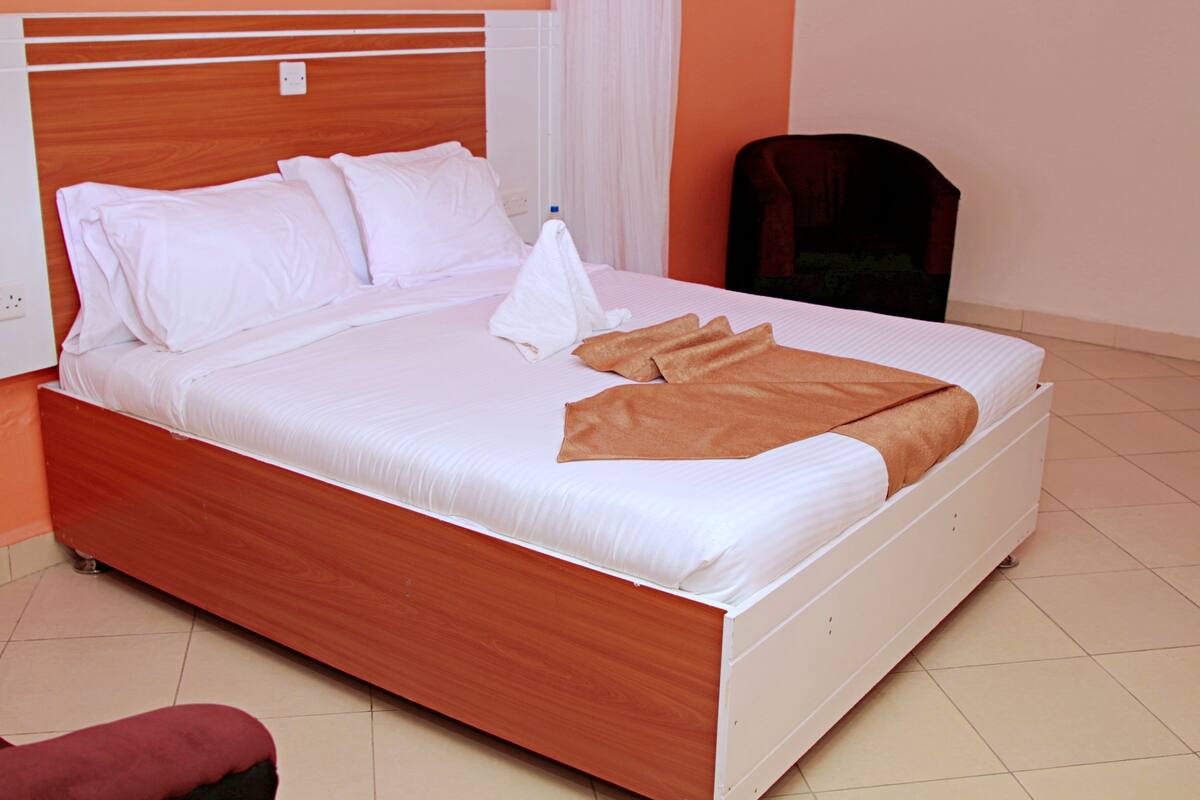 A neatly made bed with white linens and a brown blanket, a wooden headboard, and a chair in the room.