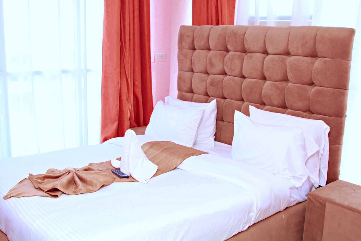A neatly made bed with white linens and a brown throw blanket, a tufted headboard, and red curtains.