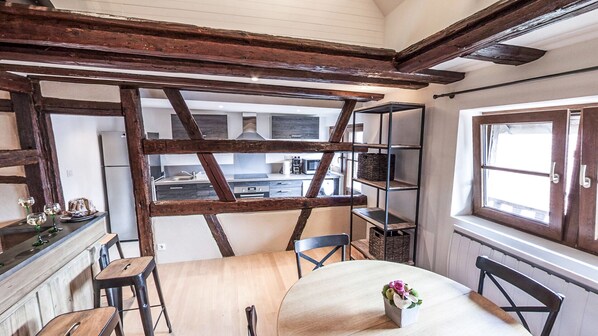 Una cocina rústica con vigas a la vista, una zona de comedor con mesa y sillas de madera, y vista a la cocina a través de un marco de madera.