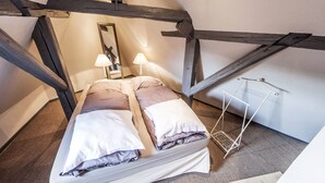 A cozy loft bedroom with exposed wooden beams, a bed with two pillows, a bedside lamp, and a hanging light fixture. - Gite 'WATCHER (Turckheim)