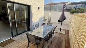 Eine Terrasse im Freien mit einem Glastisch und Stühlen, einer Holzterrasse und einer Glasschiebetür, die zu einem Innenraum führt.