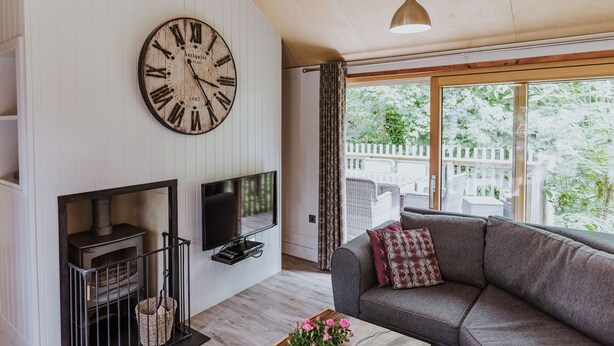 A living room with a large wall clock, a fireplace, a sofa, and a large window with a view of greenery. - Lodge 7 (Wareham)