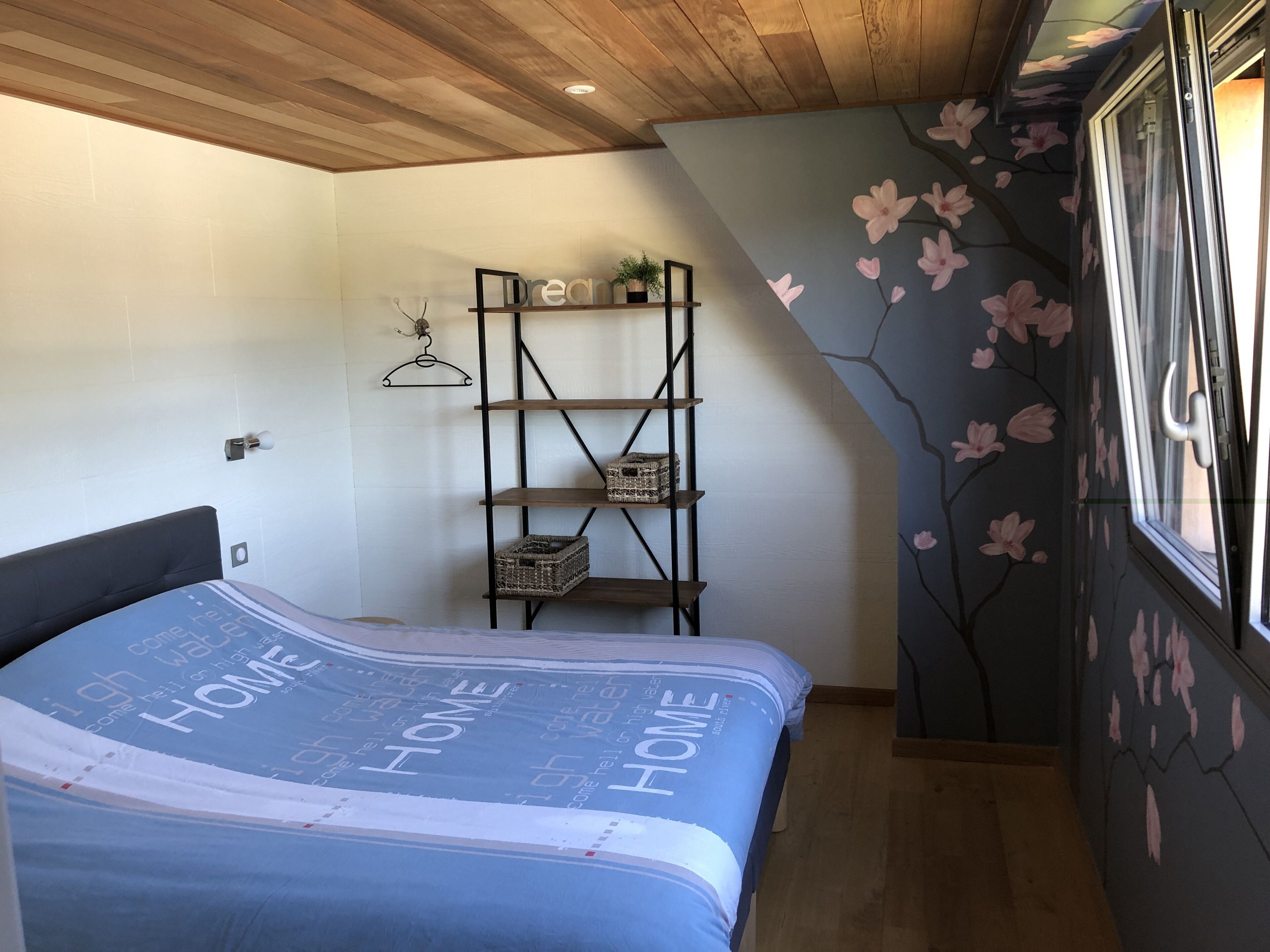 A bedroom with a bed, a wooden ceiling, a wall-mounted shelf with baskets, and a window with a floral wall design.