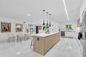 A modern kitchen with a central island, pendant lighting, and a dining area with chairs.