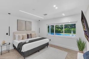 A modern bedroom with a bed, a small table, a window with a view of a swimming pool, and a potted plant.