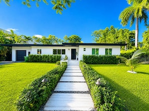 A modern two-story house with a symmetrical design, a central pathway leading to the entrance, and lush greenery surrounding the property.
