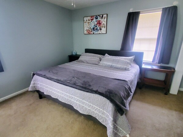 Un dormitorio con cama, mesita de noche, ventana con cortinas y una obra de arte floral en la pared.