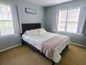 A bedroom with a bed, two windows with curtains, a picture on the wall, and a carpeted floor.