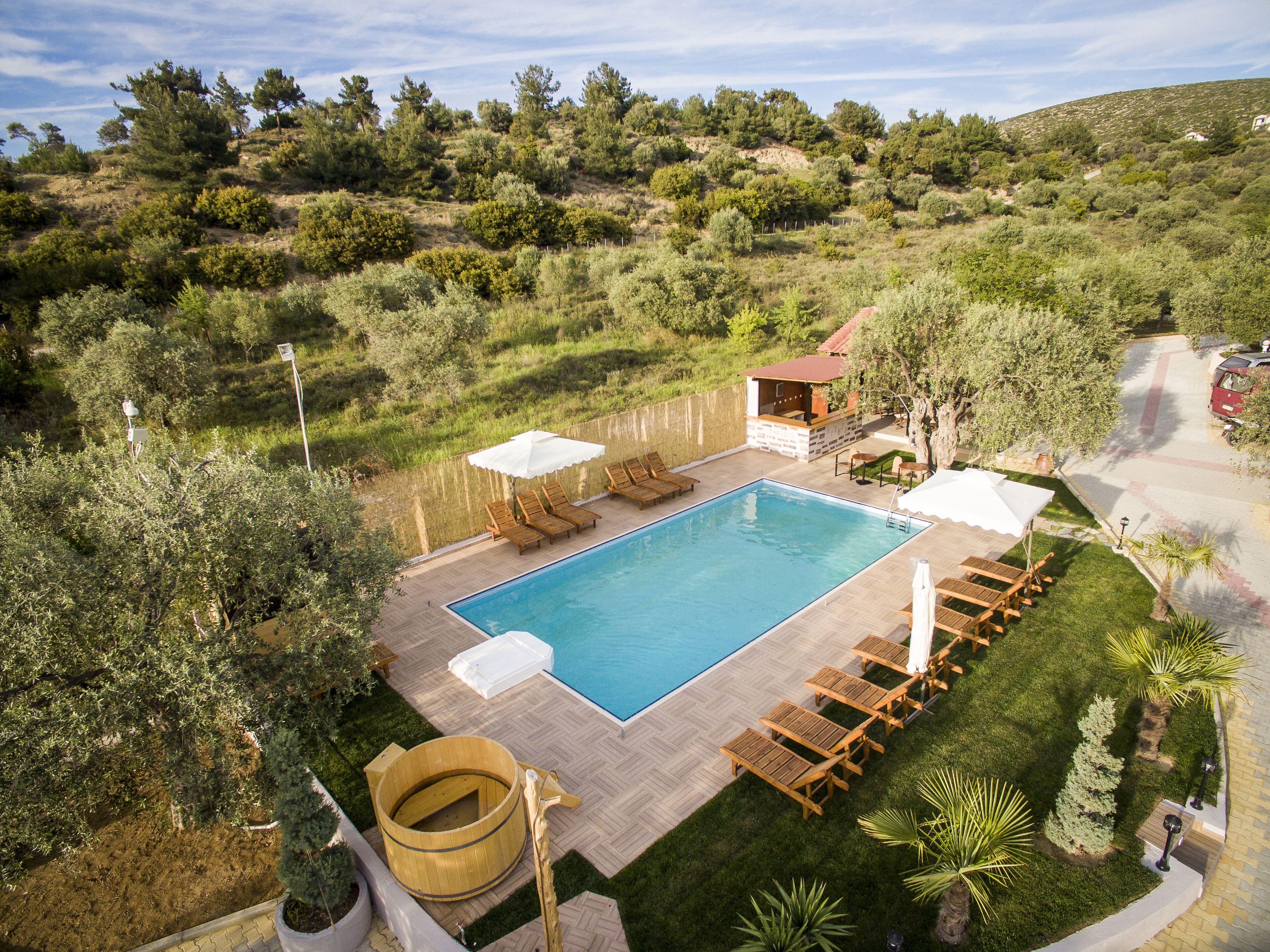 A rectangular swimming pool with a wooden deck, surrounded by lounge chairs and umbrellas, set against a backdrop of trees and hills.