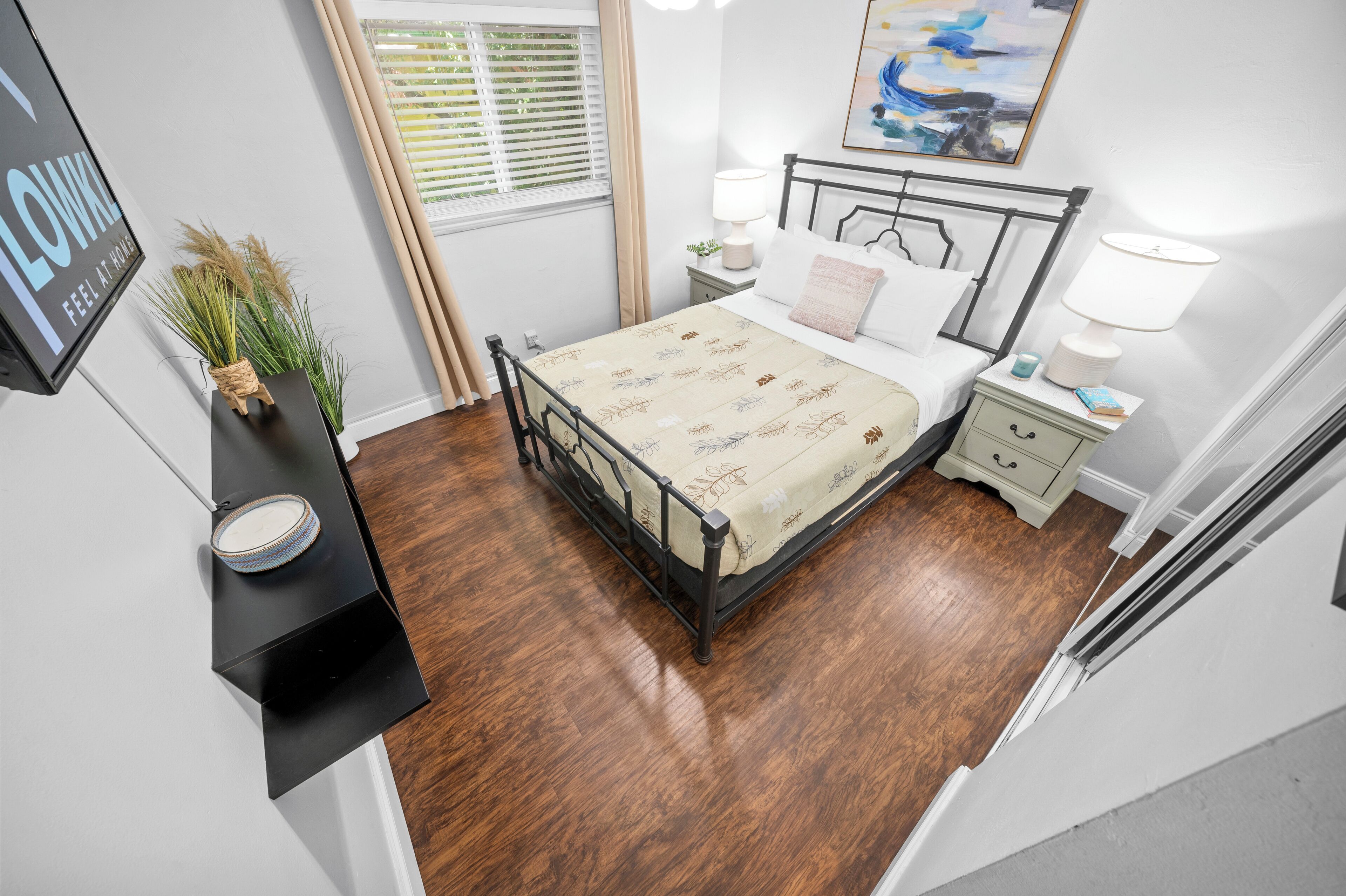 Un dormitorio con cama, mesitas de noche, una lámpara de noche, una ventana con persianas, un cuadro en la pared y una planta pequeña en un estante.
